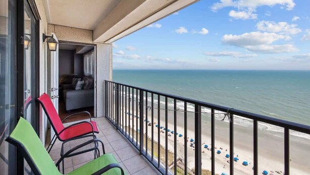 A balcony with a view of the beach, a seating area with chairs, and a glass door leading to an interior space. - Direct Oceanfront Deluxe Suite Perfect View 3BR! (Myrtle Beach)