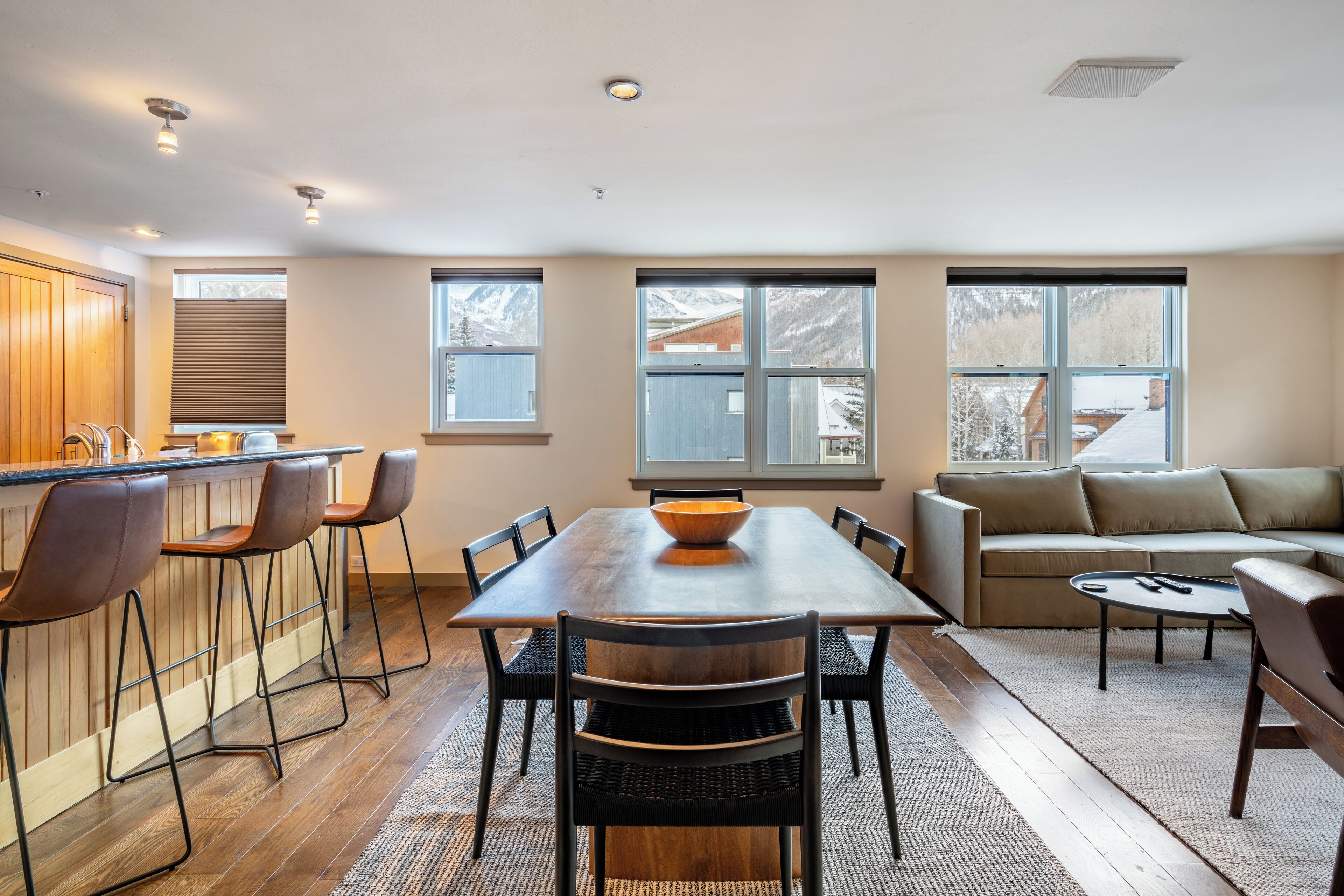 Une cuisine moderne avec un îlot central en bois, des tabourets de bar et une table à manger avec des chaises. Le salon comprend un canapé et une table basse, et de grandes fenêtres offrent une vue sur les montagnes.