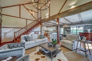 A spacious living area with a high ceiling, exposed beams, a staircase, and a kitchen in the background.