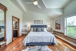 A bedroom with a large bed, wooden flooring, a ceiling fan, and a view of the outdoors.