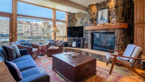 Un salón amplio con chimenea de piedra, asientos cómodos, un televisor de pantalla plana y ventanas grandes que ofrecen vistas a edificios.