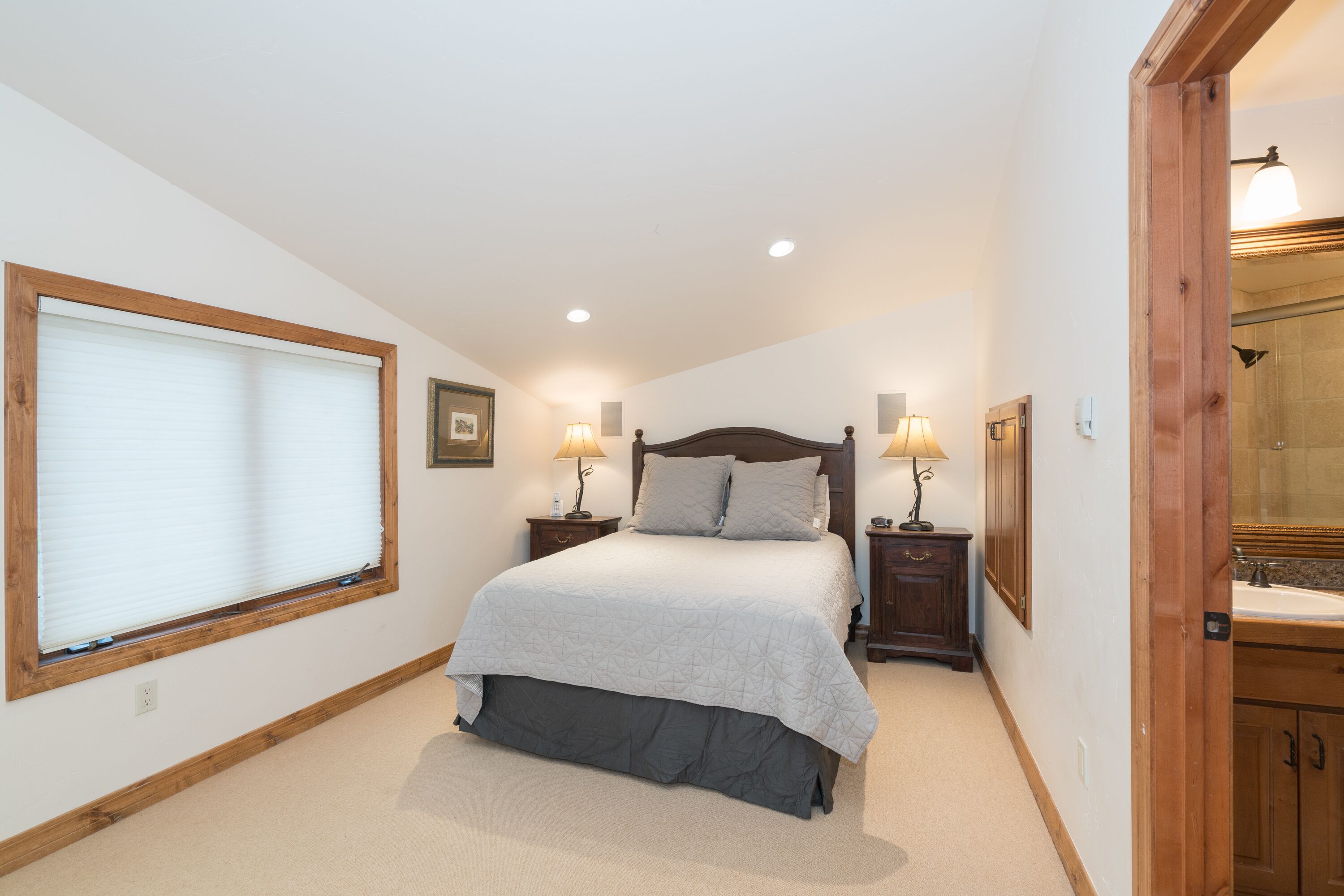 A bedroom with a bed, bedside lamps, a window with blinds, a wooden headboard, and a bathroom with a sink and mirror.