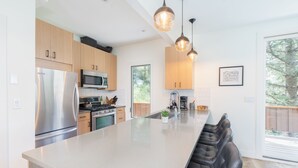 A modern kitchen with stainless steel appliances, wooden cabinets, a large island with seating, pendant lighting, and a view of the outdoors.