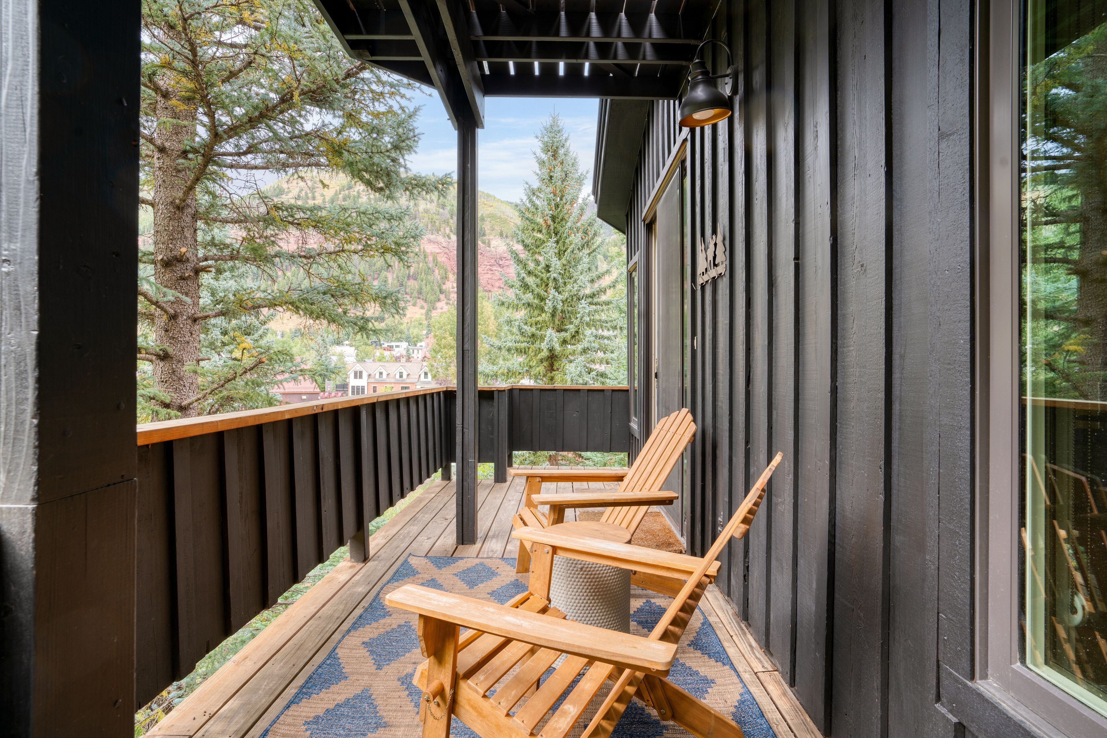 Une terrasse en bois avec deux fauteuils Adirondack, un tapis à motifs bleus et une vue sur des arbres et un bâtiment en arrière-plan.