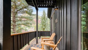 A wooden deck with two Adirondack chairs, a blue patterned rug, and a view of trees and a building in the background.