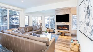 A modern living room with a sectional sofa, a fireplace, a flat-screen TV, and large windows.