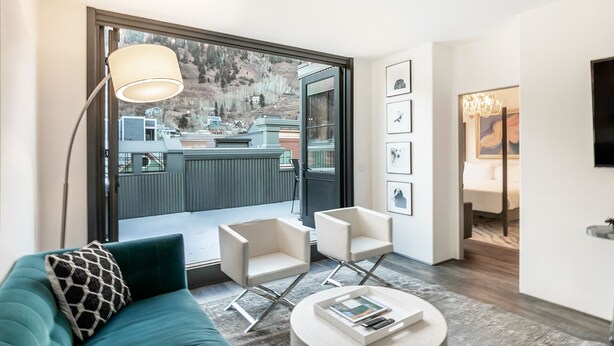 A modern living room with a teal sofa, two white armchairs, a round coffee table, and a large window offering a view of a mountain and a building.