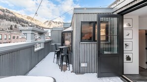 A modern outdoor balcony with a metal railing, a small seating area with chairs, and a door leading to an interior space.