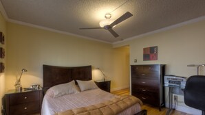 Un dormitorio con cama, mesitas de noche, escritorio, silla y ventilador de techo.