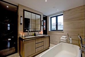 A modern bathroom with a large mirror, dual sinks, a bathtub, and a window providing natural light. - 3-bed Penthouse (London)