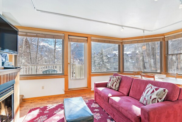 A cozy living room with a red sofa, a wooden bench, and large windows offering a view of snow-covered mountains.