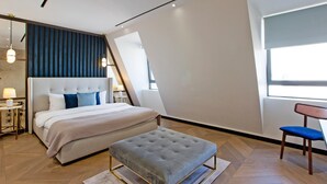 Une chambre à coucher moderne avec un grand lit, un pouf capitonné, une chaise et une fenêtre avec des stores.