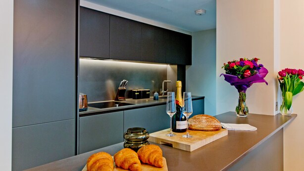 Una cocina moderna con armarios oscuros, encimera de madera, una botella de champán, dos copas, un pan y un jarrón con flores.