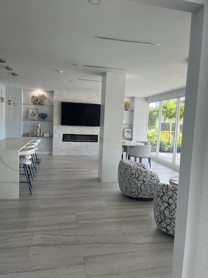 A modern kitchen with a built-in TV, a dining area with bar stools, and a seating area with patterned ottomans.