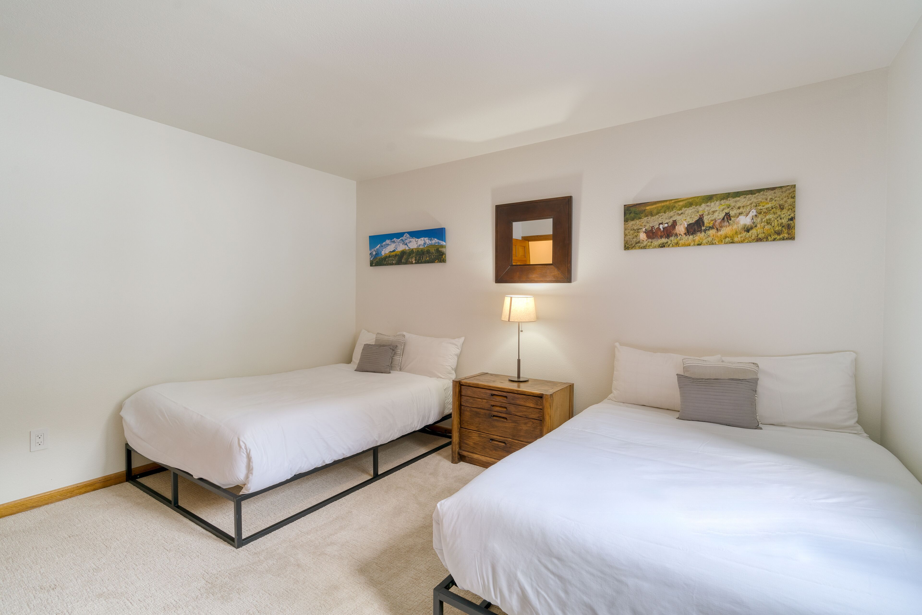 A room with two single beds, a wooden nightstand with a lamp, and two wall-mounted paintings.