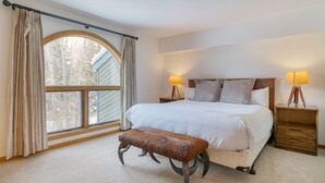 A bedroom with a large bed, two bedside tables with lamps, a bench, and a window with curtains. - River-View Condo • Walk to Lift #8 + Gondola + Downtown | Riverside Condos C102 (Telluride)