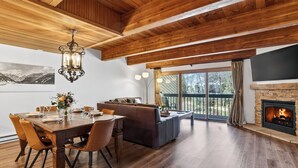 A spacious room with a wooden ceiling, a fireplace, a dining area with a table and chairs, and a large window with a view of the outdoors. - River-View Condo • Walk to Lift #8 + Gondola + Downtown | Riverside Condos C102 (Telluride)