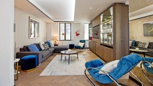 Un salon moderne avec un canapé, un pouf et des fauteuils bleus, un coin repas avec une table et des chaises, et une cuisine équipée d’un réfrigérateur et d’étagères.
