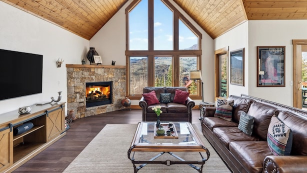 A living room with a stone fireplace, large windows, a television, and a leather sofa. - Russell Home by AvantStay | Expansive Deck, Stunning Views & Hot Tub! (Telluride)