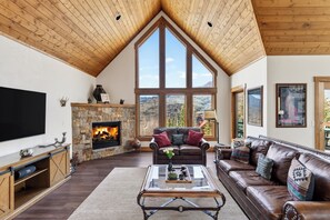 A living room with a stone fireplace, large windows, a television, and a leather sofa. - Russell Home by AvantStay | Expansive Deck, Stunning Views & Hot Tub! (Telluride)