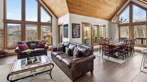 A spacious living room with a high ceiling, large windows, a leather sofa, a dining table, and chairs. - Russell Home by AvantStay | Expansive Deck, Stunning Views & Hot Tub! (Telluride)