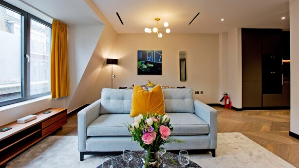 Ein modernes Wohnzimmer mit einer Couch, einem Couchtisch mit Vase und Gläsern, einem großen Fenster mit Vorhängen und einer Deckenleuchte.