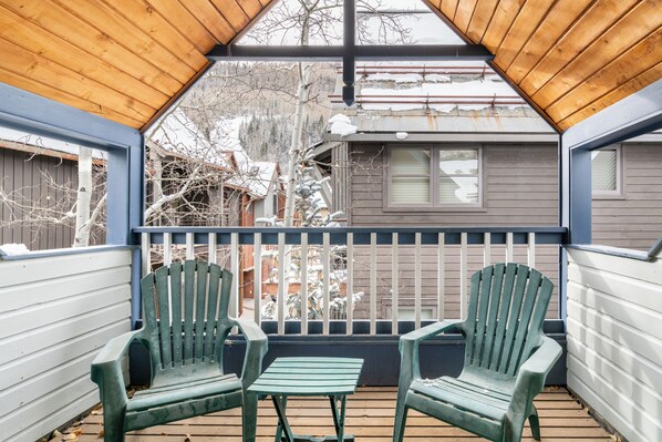 Un porche cubierto con dos sillas Adirondack verdes y una mesita, con vistas a una casa con barandilla metálica y árboles cubiertos de nieve.
