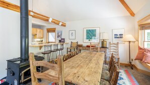 Une pièce chaleureuse avec une grande table à manger en bois, un poêle en bois noir, des chaises en bois, un coin fenêtre avec un coussin, et un espace cuisine doté de meubles en bois et d’un plan de travail.