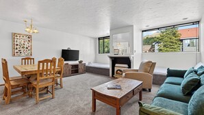 A living room with a wooden dining table, chairs, a sofa, a fireplace, a TV, and a large window with a view of trees and buildings. - Telluride Lodge 311 by AvantStay | Close to Slopes & Town! (Telluride)