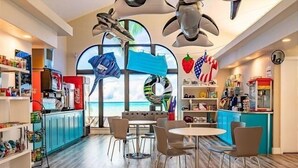 Una habitación luminosa con un amplio ventanal con vista a una playa, que cuenta con una zona de cocina con armarios azules, una mesa de comedor con sillas y varios electrodomésticos.