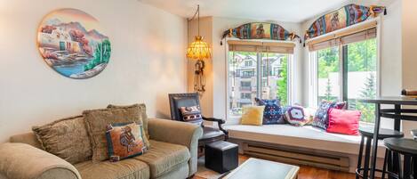 A cozy living room with a sofa, a coffee table, and a window seat with colorful cushions.