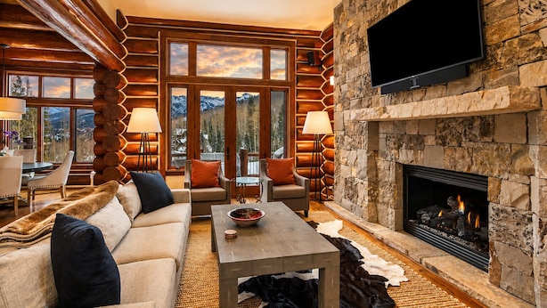 L’intérieur d’une cabane en rondins, avec une cheminée en pierre, un téléviseur fixé au mur et une vue sur les montagnes par de grandes fenêtres.