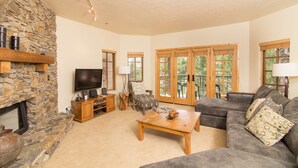 A living room with a stone fireplace, a flat-screen TV, a wooden coffee table, a gray sofa, and a large window with a view of trees. - Westermere 311 - 2 by AvantStay | Just Steps From Ski Area w/ Great Views! (Telluride)