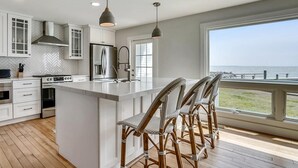 A modern kitchen with white cabinetry, a large island with bar stools, stainless steel appliances, and a view of the ocean through a large window. - Tilghman Waterfront | Indoor Pool | Private Dock | Pool House (Tilghman)