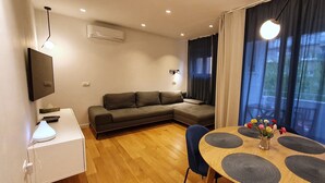 A modern living room with a sofa, a dining table with placemats, a TV mounted on the wall, and a window with curtains. - Apartment Freyja (Split)