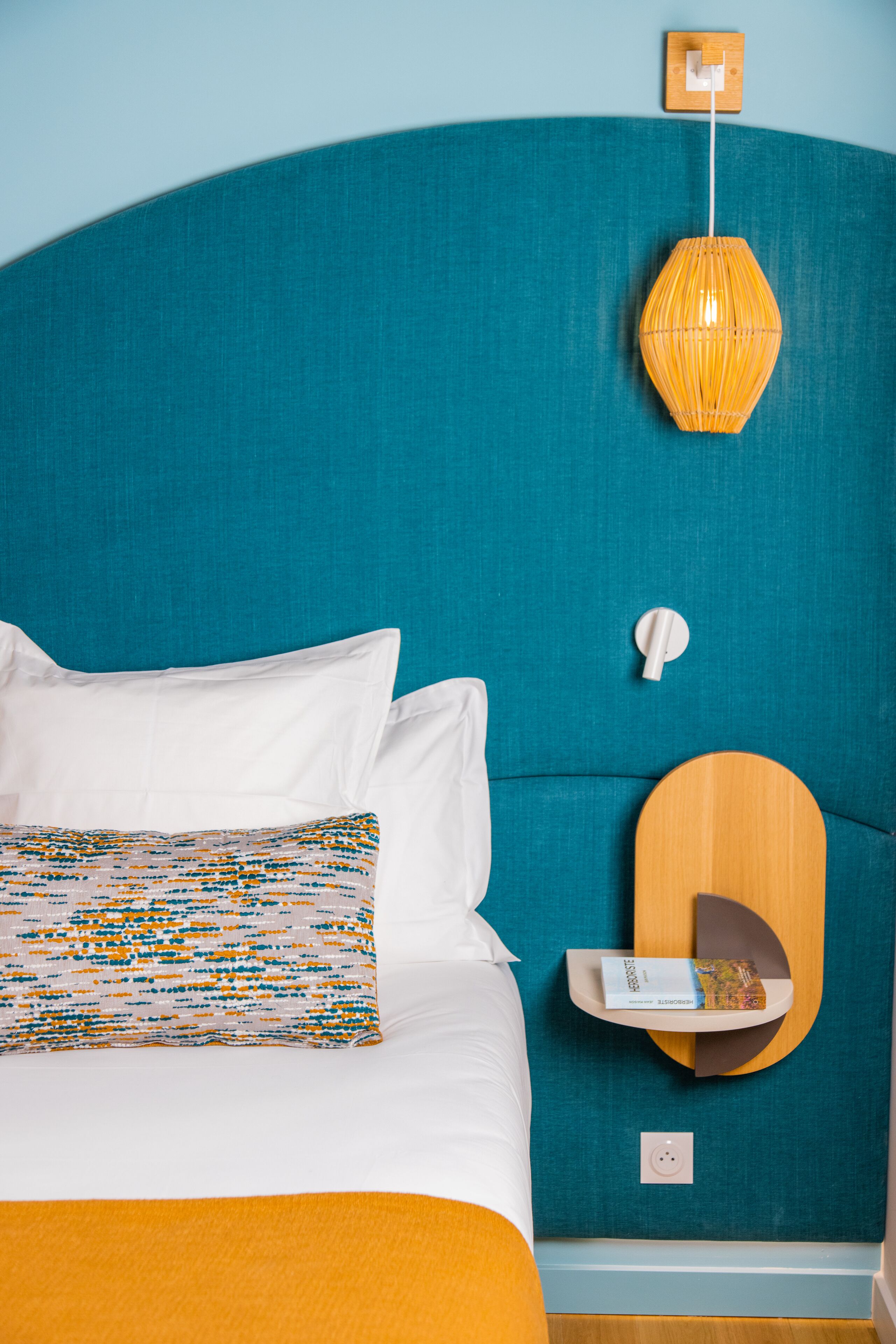 A neatly made bed with white and patterned pillows, a teal headboard, a wooden bedside table with a book, and a wall-mounted lamp.