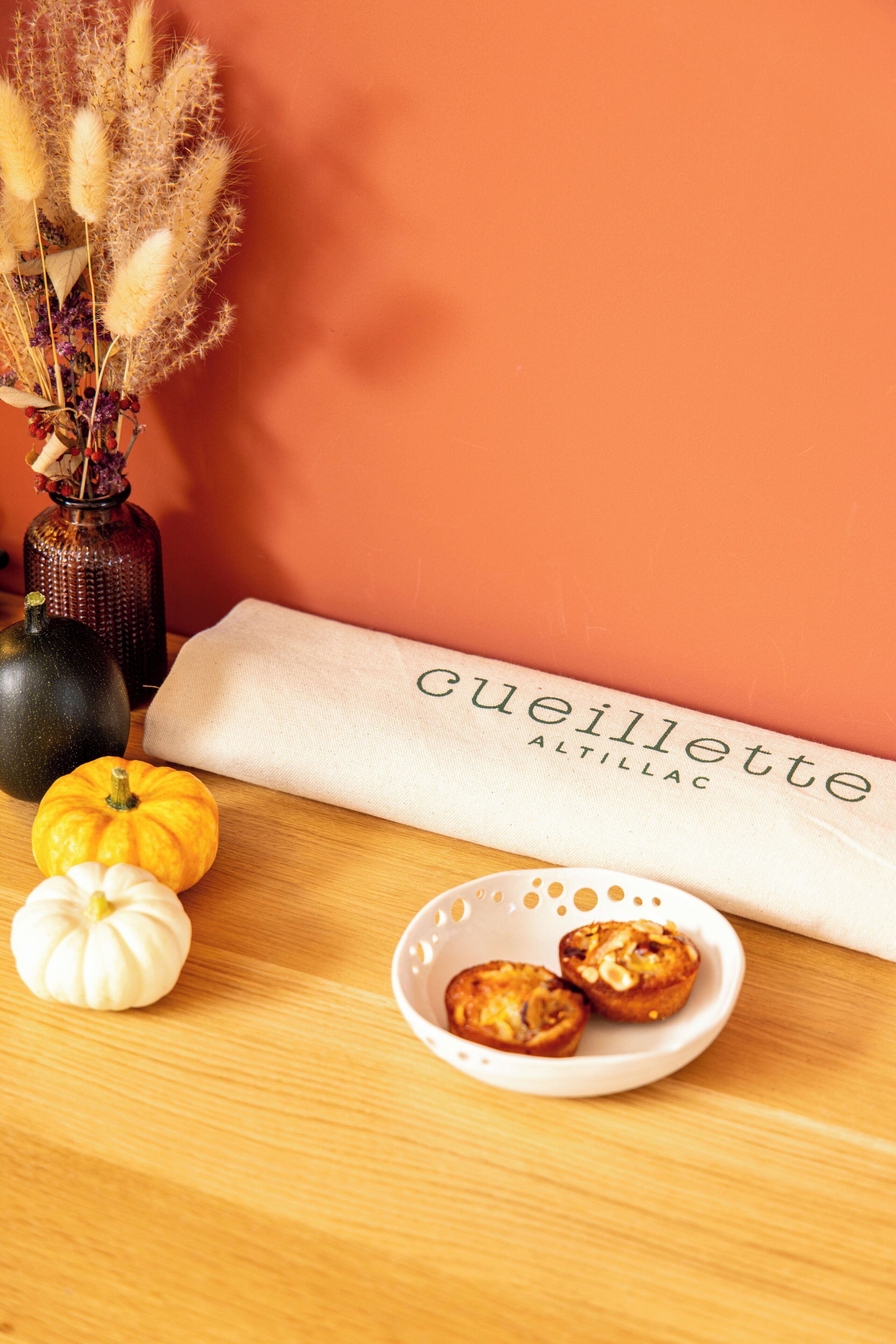A table with a vase of dried flowers, a bowl of muffins, and a small pumpkin.