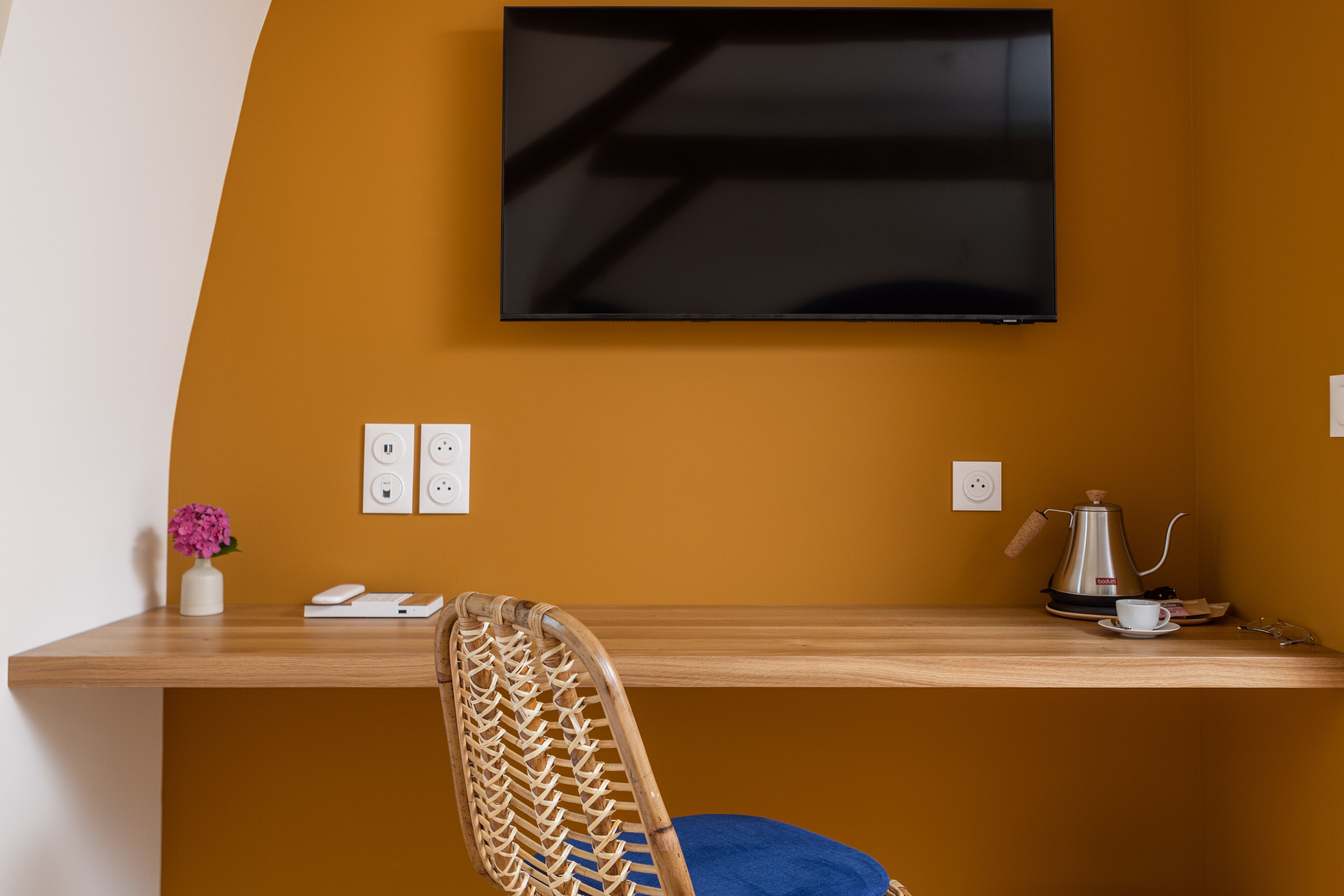 A room with a wooden desk, a chair, a television mounted on the wall, and a coffee maker on the desk.