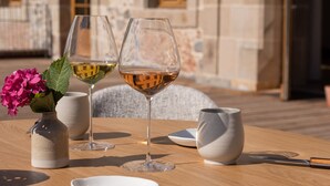 Una mesa de madera redonda con dos copas de vino, un jarrón blanco con flores rosadas y una jarra blanca.