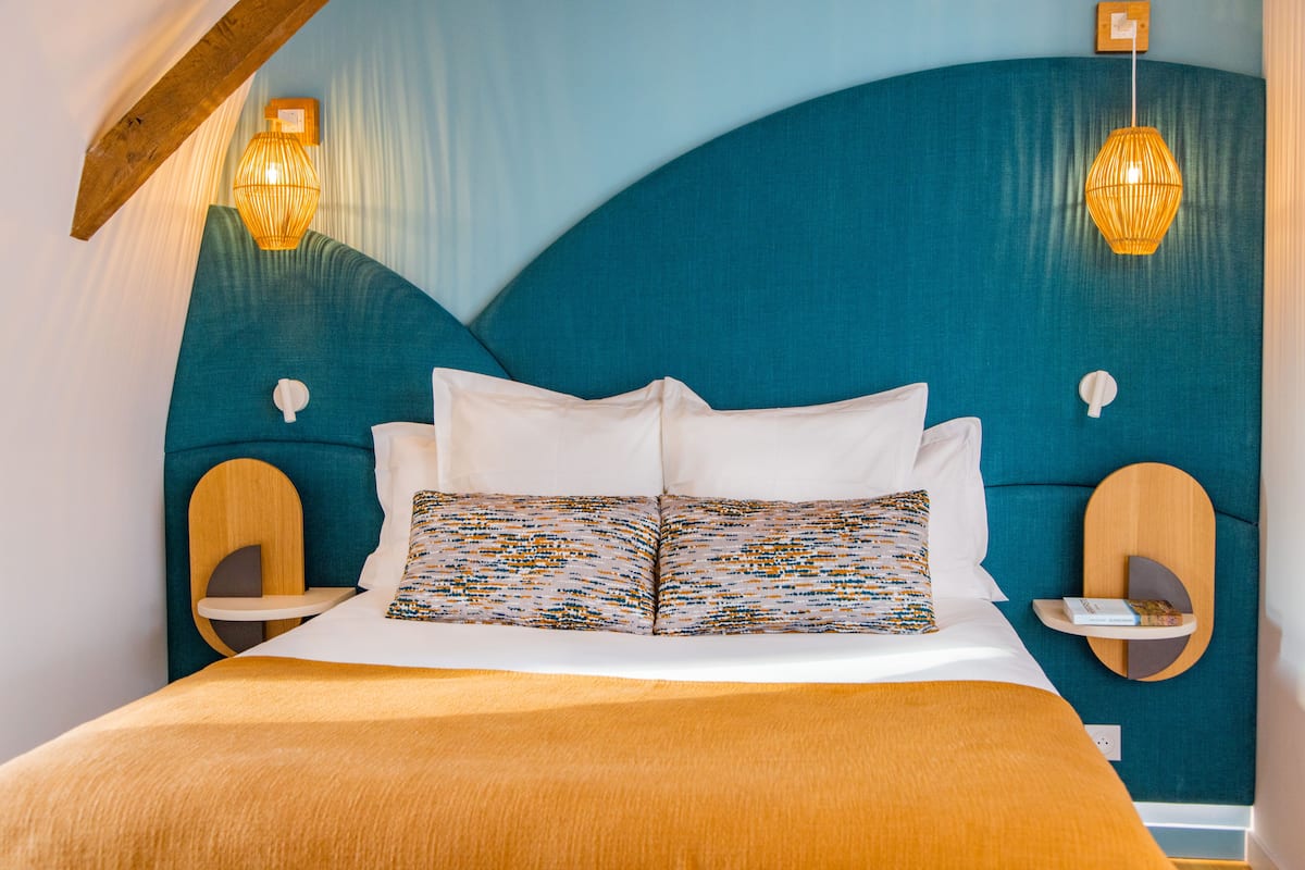 Un lit avec des oreillers blancs et à motifs, une tête de lit de couleur turquoise et deux lampes fixées au mur.