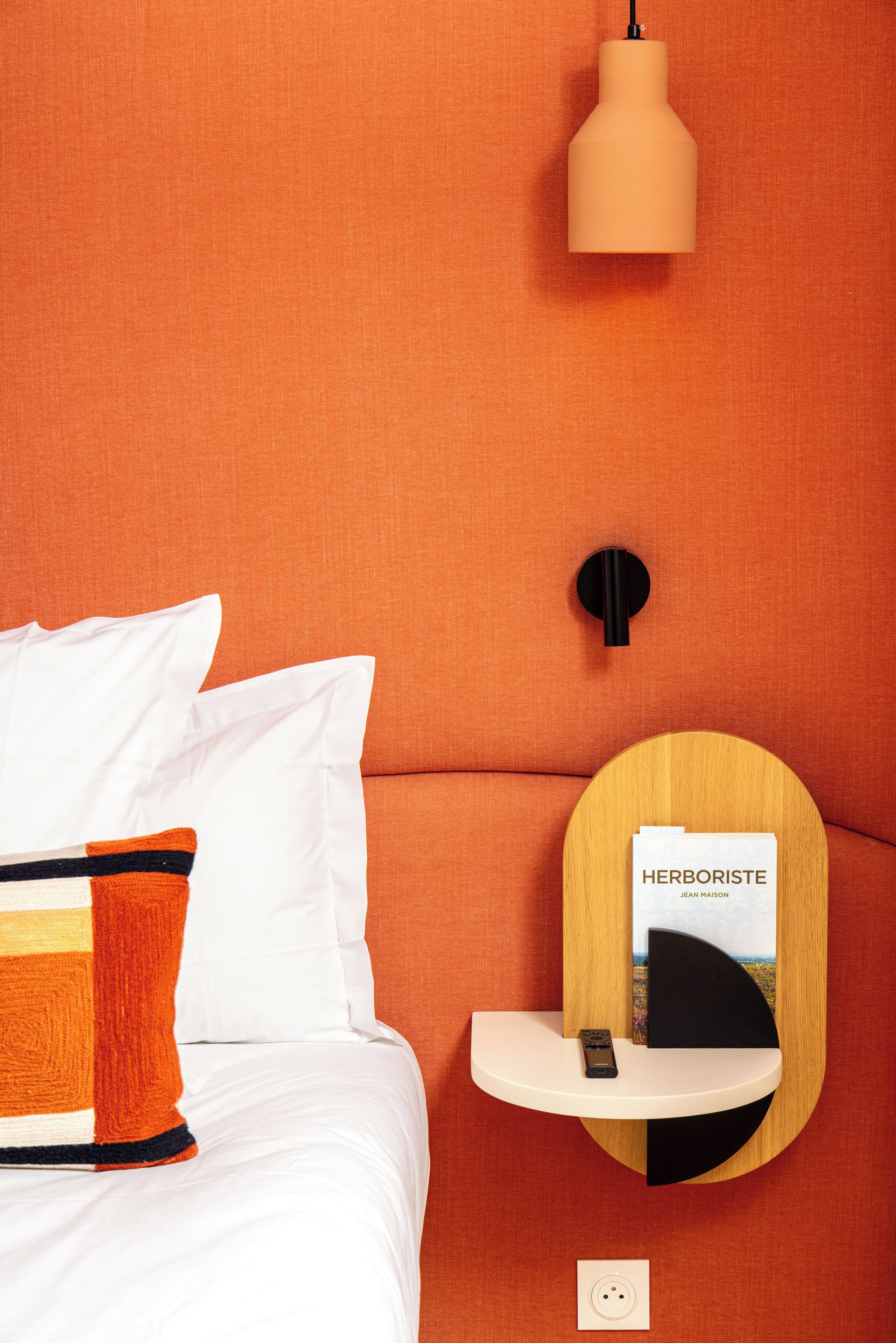 A bed with white bedding and a colorful pillow, a round wooden shelf with a book titled "HERBORISTE" and a black object, and a pendant light above the shelf.