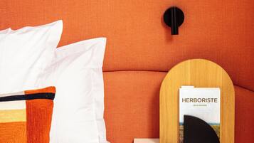 A bed with white bedding and a colorful pillow, a round wooden shelf with a book titled "HERBORISTE" and a black object, and a pendant light above the shelf.