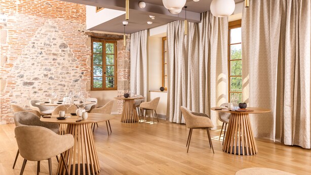 Un comedor moderno con mesas y sillas de madera, una pared de piedra y grandes ventanas con cortinas.