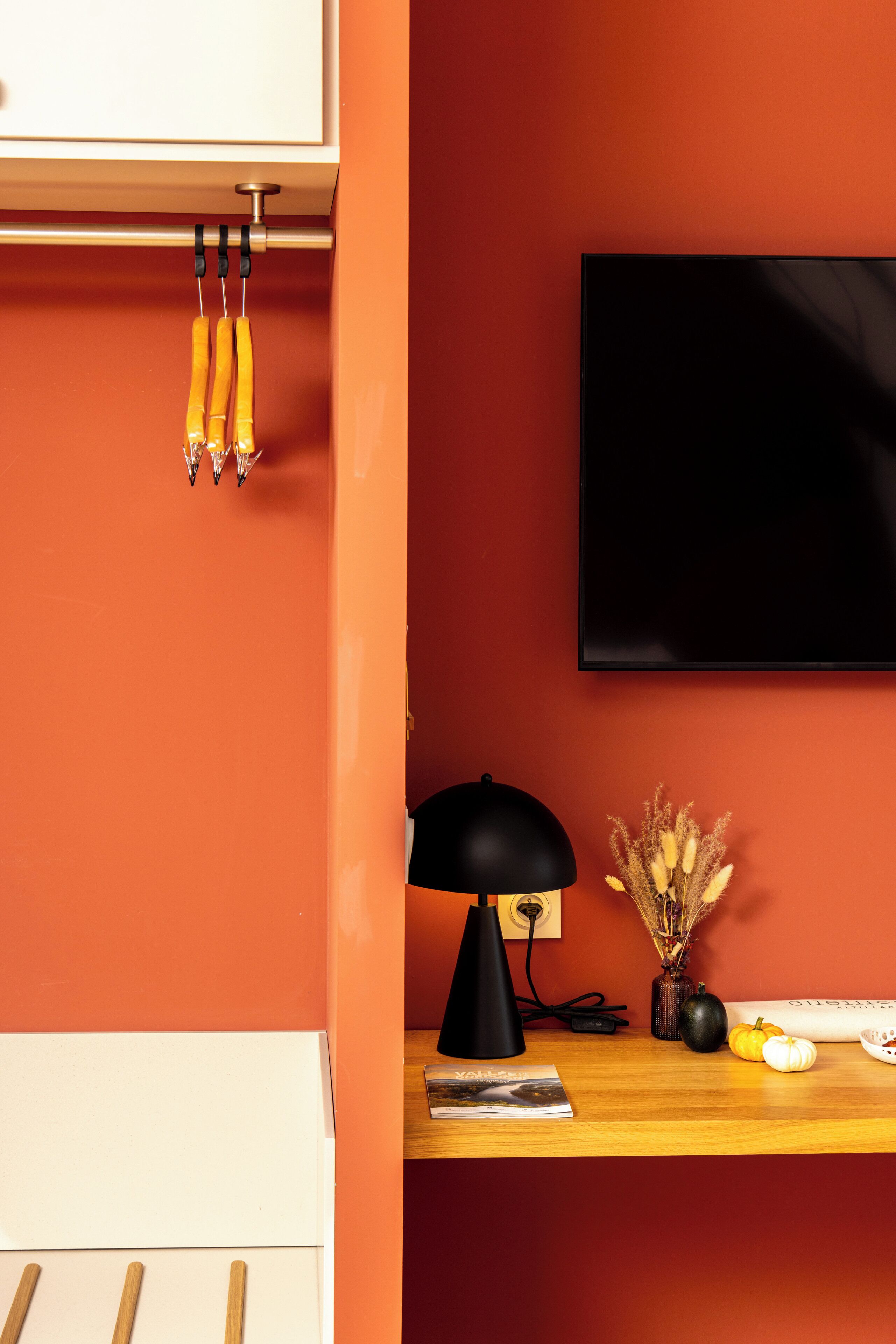 Una habitación con una pared roja, un televisor de pantalla plana sobre un escritorio de madera, una lámpara de escritorio negra y un florero con plantas secas.