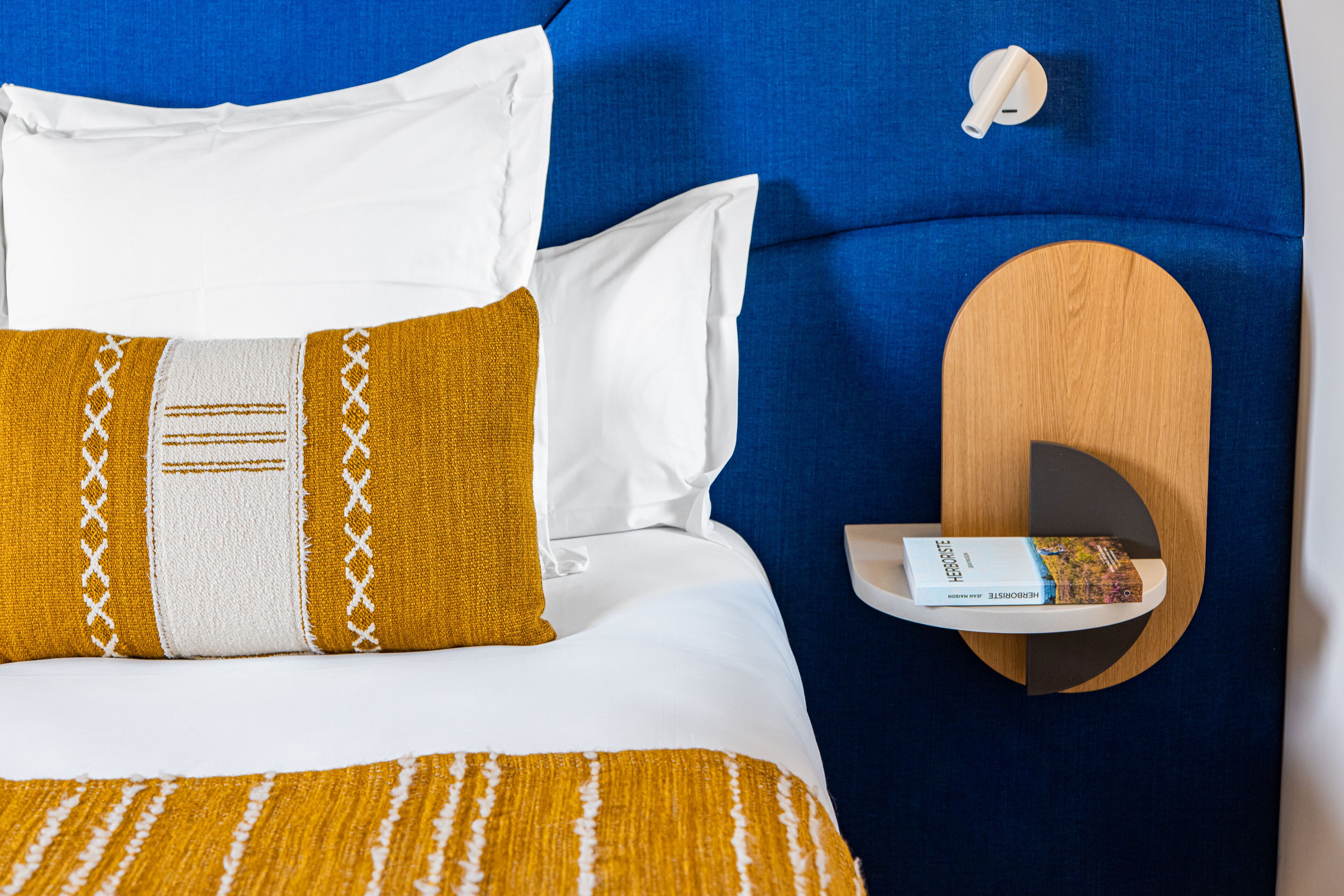 A neatly made bed with a patterned blanket, a white pillow, and a mustard-colored pillow. There is a wooden shelf with a book on it.