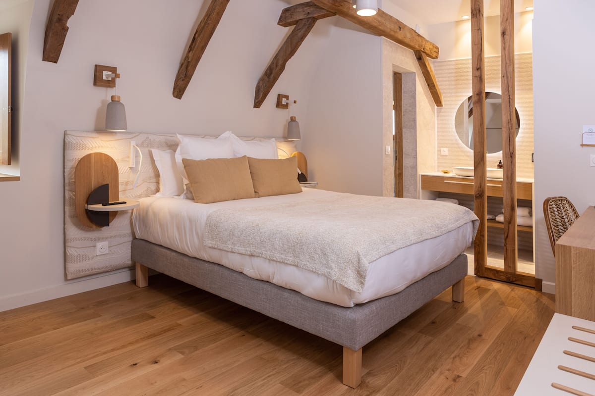 Une chambre à coucher avec un lit, des poutres en bois, une table de chevet avec une lampe, un miroir et une salle de bain visible à travers une porte ouverte.