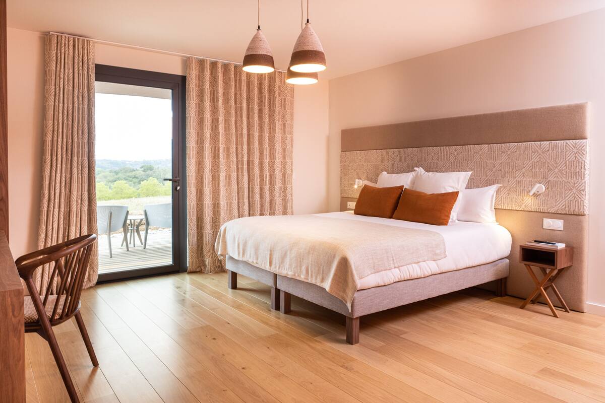 Une chambre à coucher comprenant un lit, une chaise en bois, une table de chevet et une vue sur un espace extérieur, accessible par une porte vitrée.