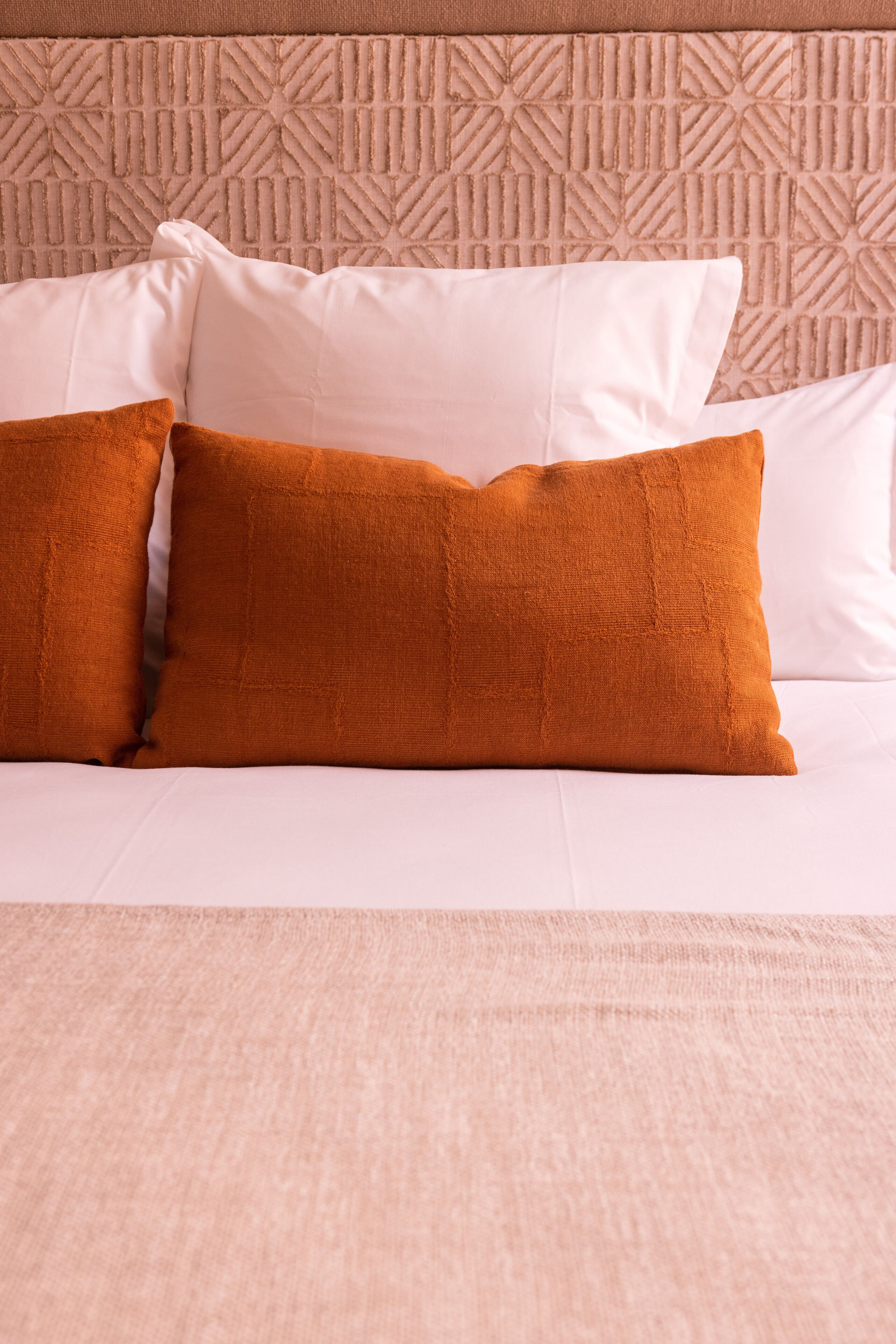 Una cama con almohadas blancas y anaranjadas, un cabecero color beige con diseño geométrico y una colcha de tonos claros.