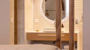Un dormitorio con cama, vigas de madera, un baño con espejo redondo y lavamanos.