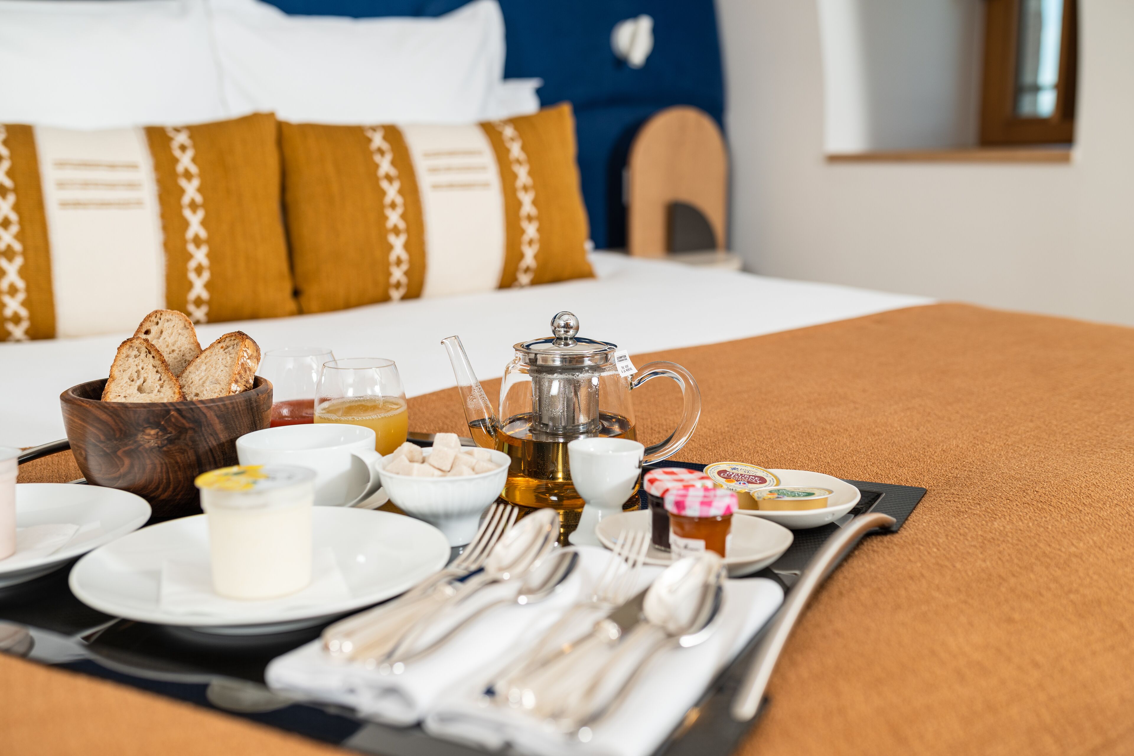 Un plateau de petit-déjeuner avec du pain, du beurre, de la confiture, du thé et du miel, posé sur un lit recouvert d’une couverture marron et agrémenté d’oreillers à motifs.